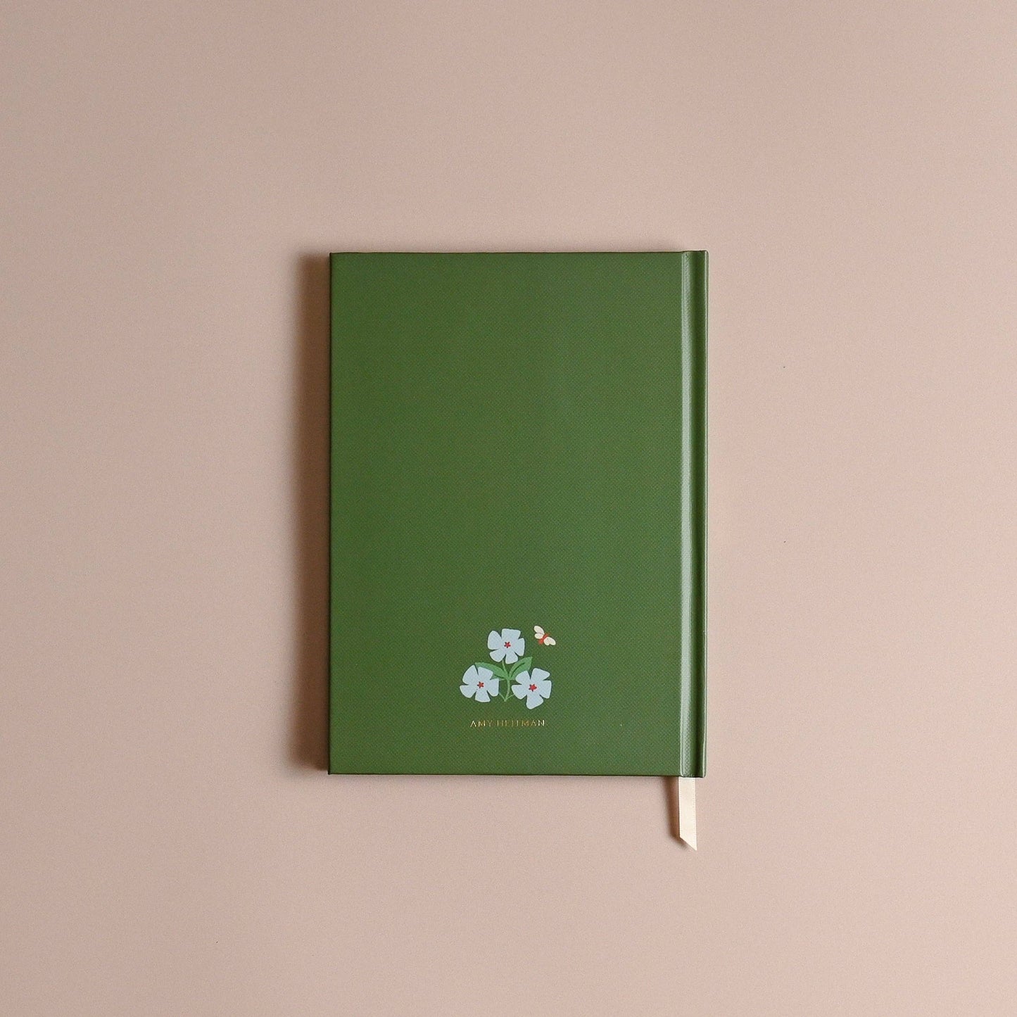 A green hardcover notebook with a small white floral and bird illustration near the bottom, photographed against a plain dusty pink background.