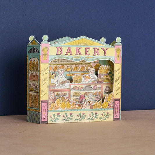 A colorful 3D papercraft model of a vintage bakery storefront, displayed on a light wooden surface against a dark blue background.