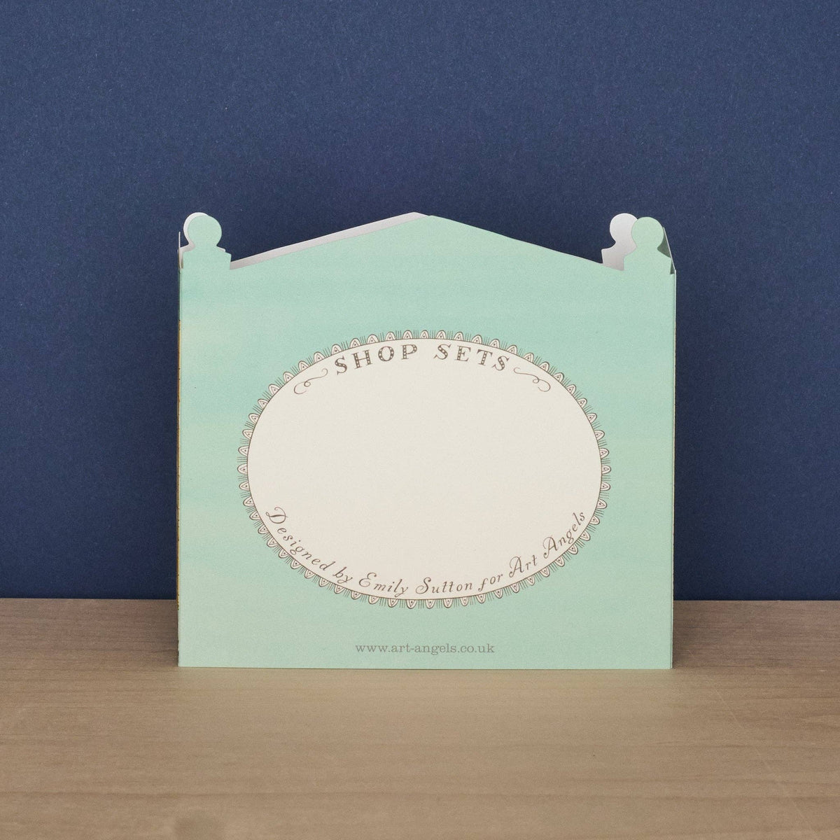 A mint green paper display stand or shop set card with a white oval center, photographed standing on a light wood surface against a dark blue background. The front reads 'SHOP SETS' with decorative script text and a small website at the bottom.