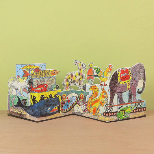 A colorful 3D pop-up cardboard animal storybook displayed on a light wooden surface against a pale yellow-green background. The pop-up features various animals including an elephant, rabbit, rooster, squirrel, whale, lion, and fish, each with vibrant painted details and traditional patterns.