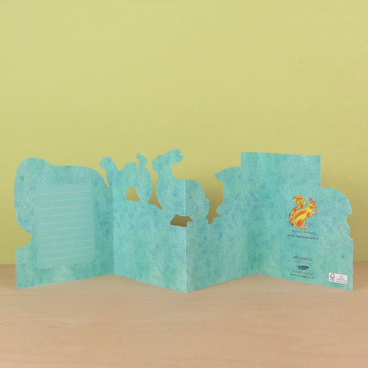 A handmade paper pop-up card featuring a light blue textured background and cutout animal silhouettes, displayed on a light wood surface against a pale yellow-green background.
