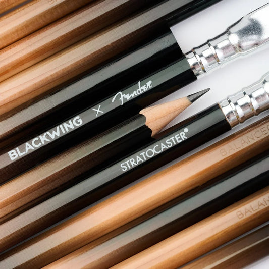 Close-up of several Fender Blackwing Stratocaster pencils arranged diagonally, showing black ferrules, silver metal tips, and natural wood barrels with white printed branding.