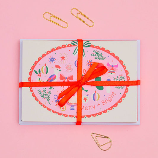 A square greeting card with a colorful Christmas-themed oval design and the words 'Merry & Bright' at the bottom, wrapped with a bright red ribbon and tied in a bow, on a solid pink background with gold paperclips.
