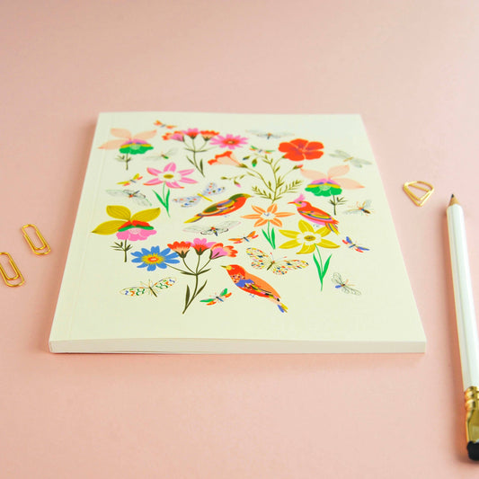 A cream-colored notebook with a colorful floral and bird illustration, styled on a dusty pink background with gold paperclips and a white pencil.