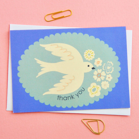Greeting card with a dove and flowers on a blue background, surrounded by paper clips on a pink background.