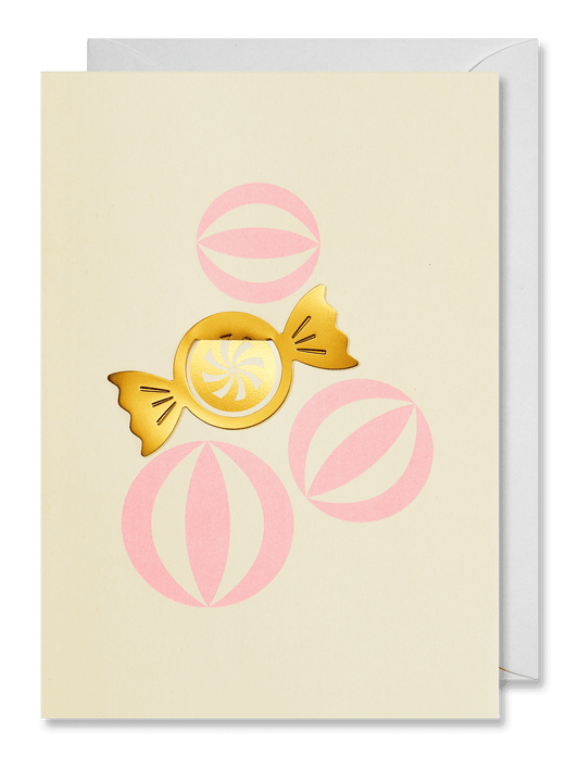 A cream-colored greeting card featuring a gold foil wrapped candy and pink circular patterns, shown with a white envelope behind it on a dark green background.