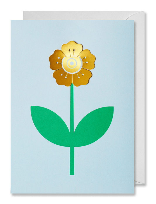 A square greeting card with a minimalist flower design, featuring a gold metallic flower head, green stem and leaves, and a pale blue background. A white envelope is partially visible behind the card against a dark green backdrop.