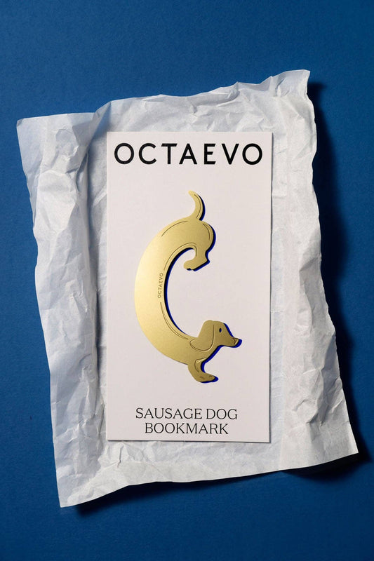 A gold sausage dog enamel bookmark by Octaevo, displayed on its white backing card and crumpled white paper against a solid blue background.