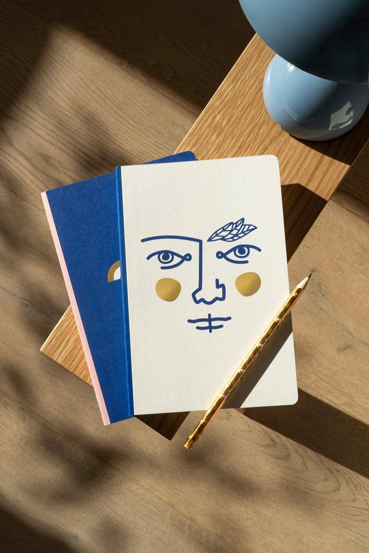 Notebook with face illustration, pen, and blue lamp on wooden surface
