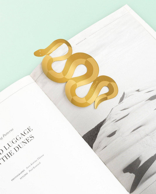 A metallic gold snake-shaped bookmark resting on open magazine pages against a pale mint background.