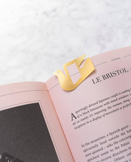 A gold metal bird-shaped bookmark clipped onto an open pink book, shown on a light marble-like surface.