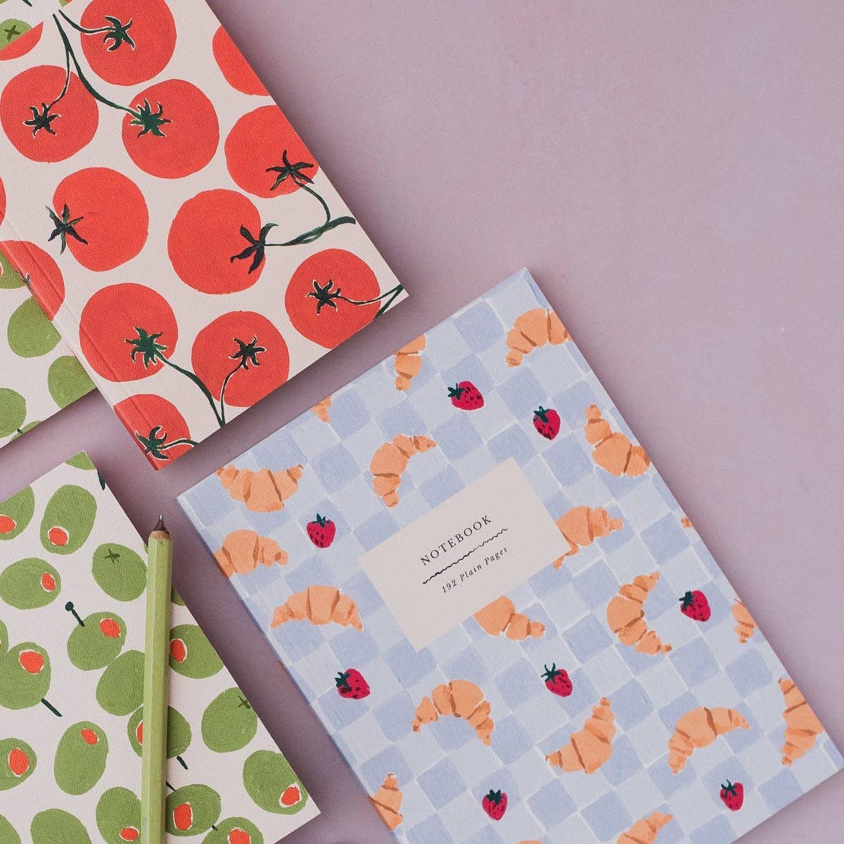 Three patterned notebooks arranged on a soft lavender background. One notebook has large red tomatoes, one has green pears, and one has orange croissants on a light blue checkered background with a white label reading 'NOTABOOK'.