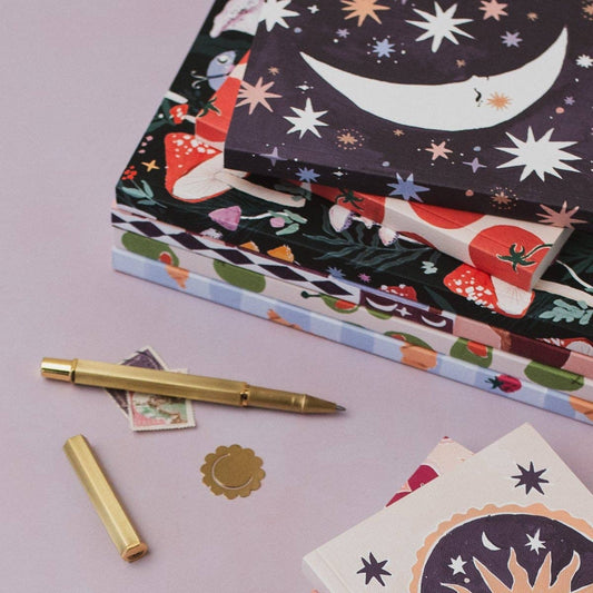 A stack of decorative notebooks with whimsical illustrated covers, including a crescent moon, stars, mushrooms, and floral patterns, arranged on a pale pink surface with a gold pen and small stationery pieces nearby.