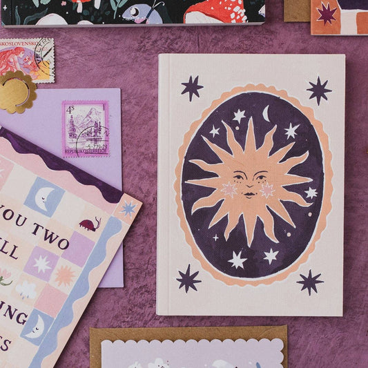 A flat-lay of stationery featuring a cream greeting card with a stylized sun and moon illustration, alongside other pastel patterned cards, envelopes, and stamps on a textured purple background.