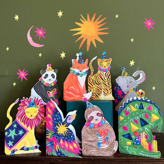 Colorful animal figurines with festive hats on a dark surface against a green background with sun and moon.