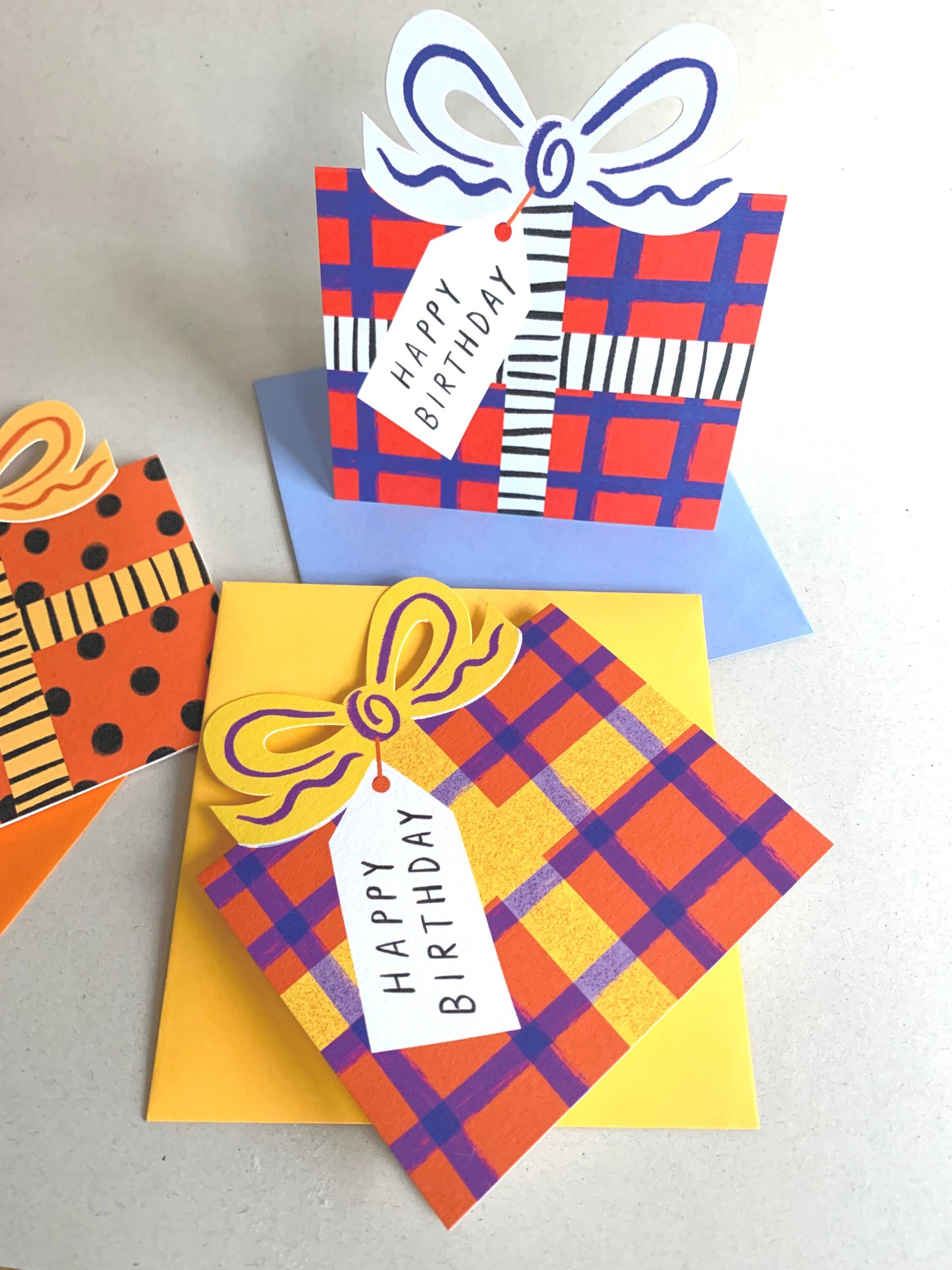 Three colorful birthday cards with plaid patterns and 'Happy Birthday' tags on a light gray background.