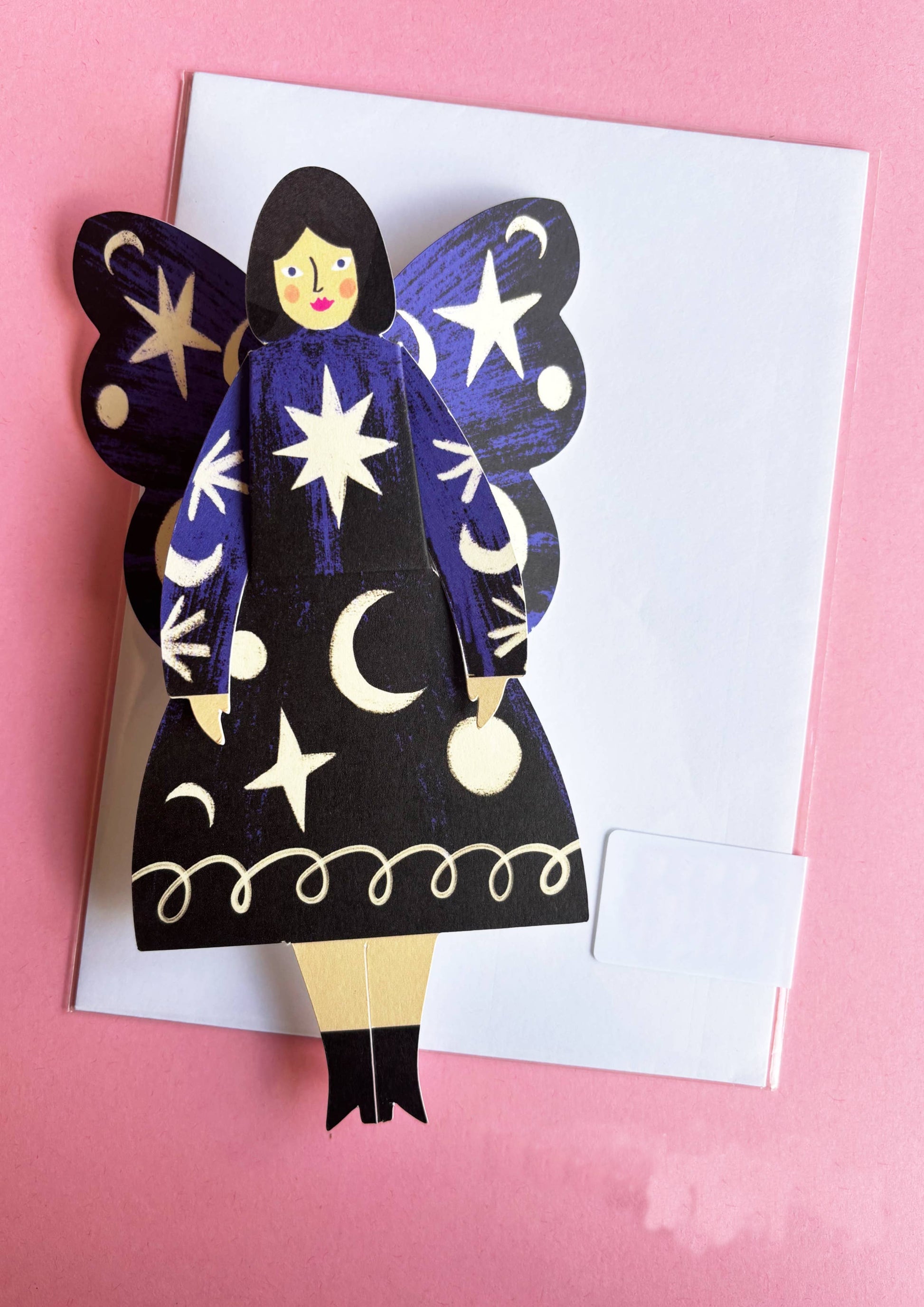 Paper cutout of a fairy with star and moon designs on a pink background