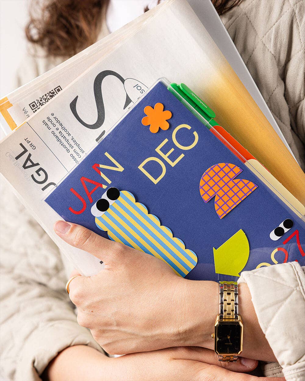 A person holding a stack of colorful books and magazines, with the frontmost book titled 'JAN DEC' in bold yellow letters on a blue cover. The person is wearing a beige quilted jacket and a gold-tone watch.