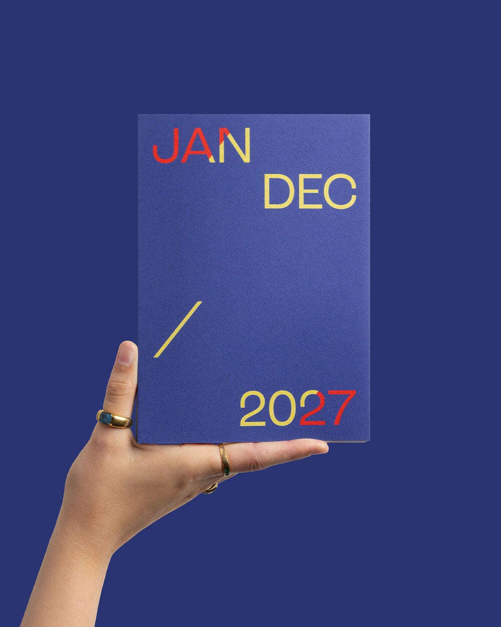 A hand holds a blue rectangular card or cover against a solid blue background. The card has the words JAN, DEC, and 2027 printed in red and yellow, with a yellow slash marking the time as one full year.