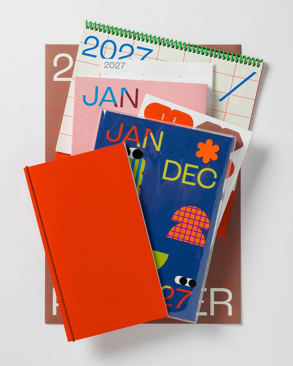 A stack of colorful printed paper goods on a light gray background, including a spiral-bound 2027 calendar, a pink January page, a brown monthly calendar, a blue December cover with abstract shapes, and a bright orange notebook or folder in front.