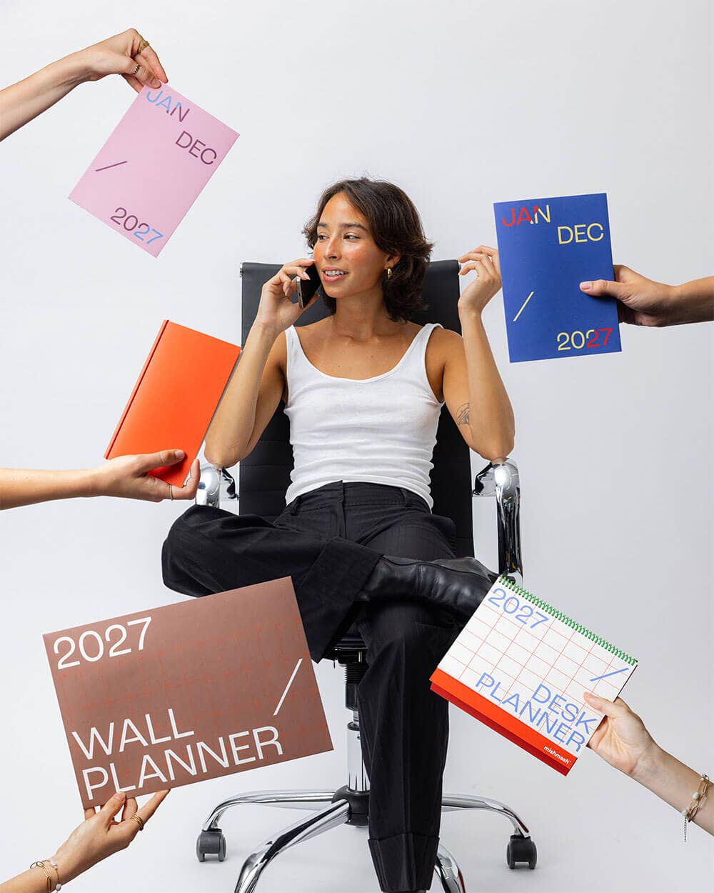 A woman in a white tank top and black pants sits in a black office chair, holding a phone to her ear while several hands around her hold up colorful 2027 planner covers and a desk planner, including pink, orange, blue, brown, and green designs, against a plain white background.