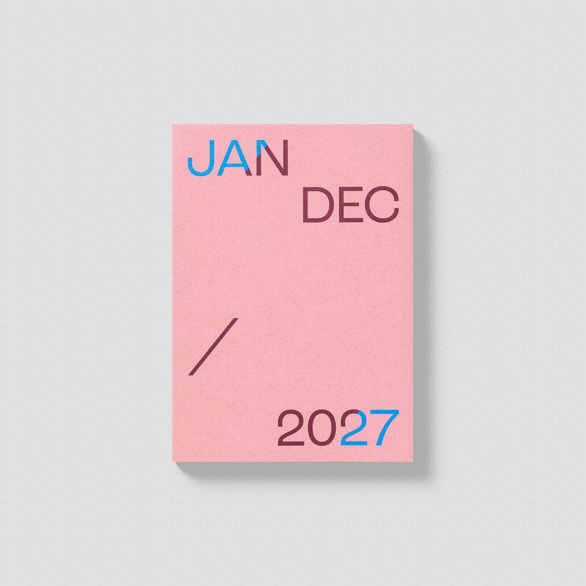 A pink rectangular card or cover with the text 'JAN DEC / 2027' in dark mauve and bright blue lettering, shown on a light gray background with a soft drop shadow.