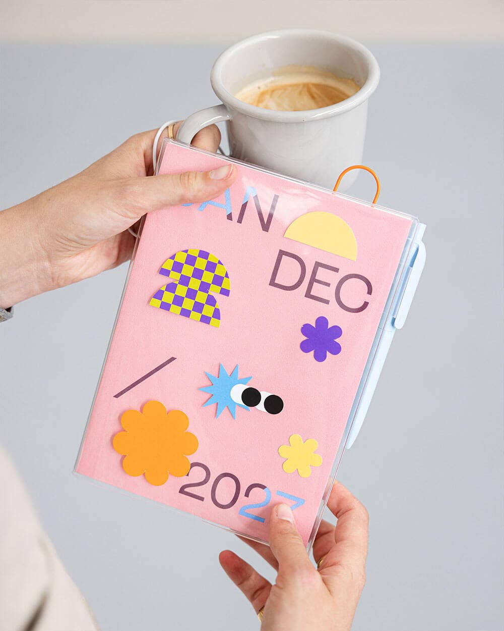 A person holds a pastel pink pocket calendar for December 2022, decorated with colorful felt shapes and text, with a white coffee mug in the background.