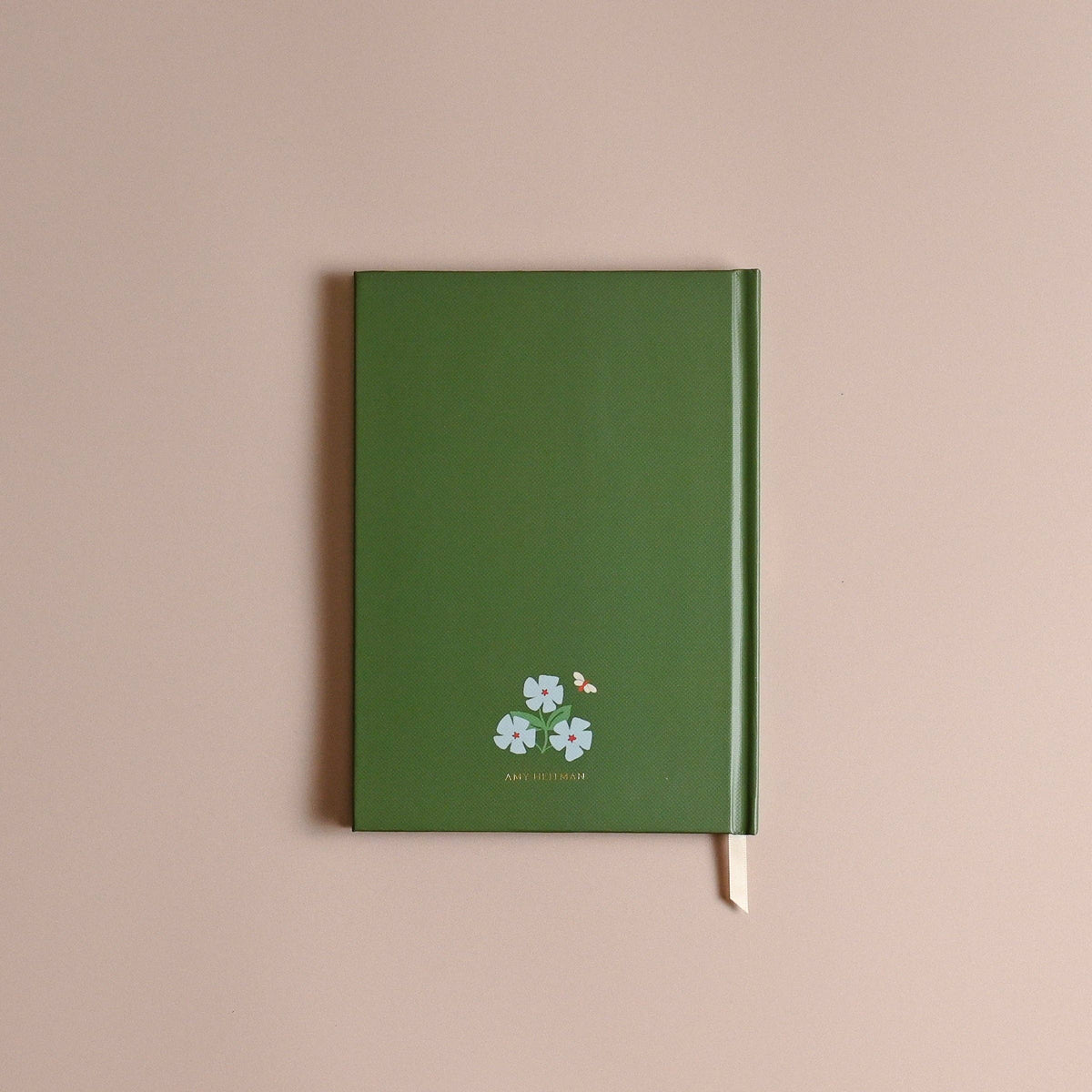 A green hardcover notebook with a small white floral and bird illustration near the bottom, photographed against a plain dusty pink background.