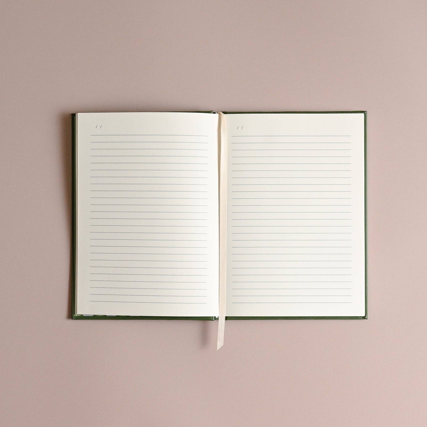 An open lined notebook with a dark green cover and a cream ribbon bookmark, displayed flat against a solid dusty pink background.