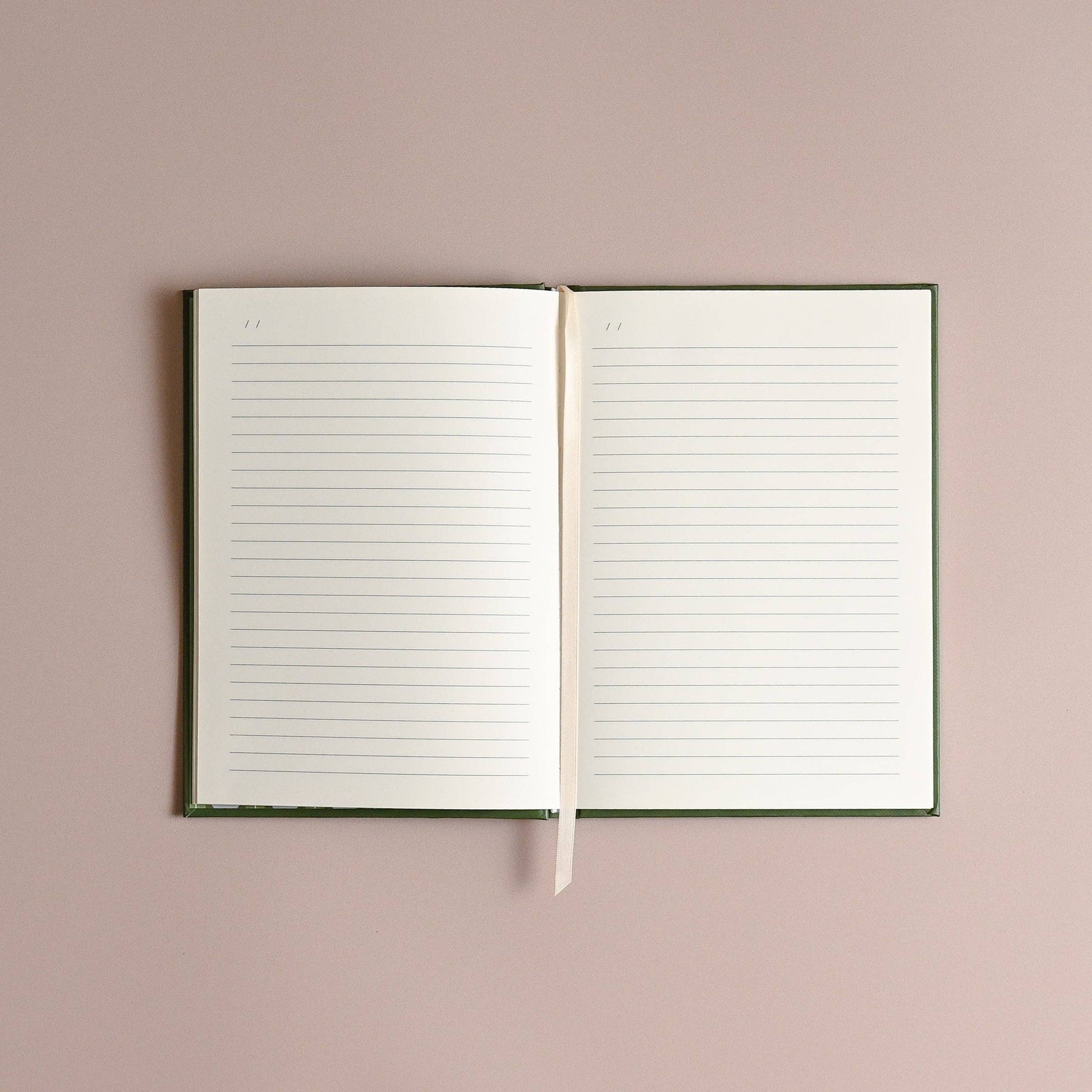 An open lined notebook with a dark green cover and a cream ribbon bookmark, displayed flat against a solid dusty pink background.