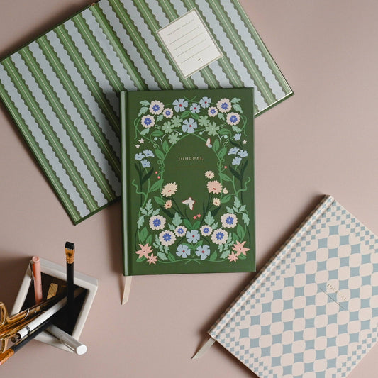 A green hardcover journal with a floral border and the word 'BIRTHDAY' on the cover, styled on a dusty pink background with other patterned notebooks and stationery.