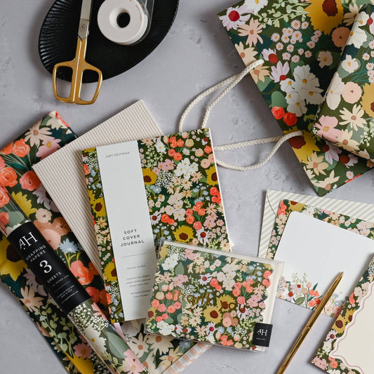 A coordinated flat-lay of floral stationery and gift items on a light gray surface, including patterned paper bags, envelopes, a notebook, a pen, a notebook roll, and a black plate with a gold-handled scissors and tape.
