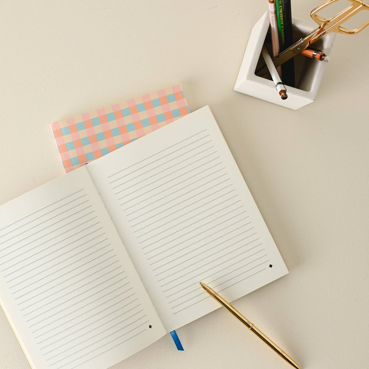 An open lined notebook on a light beige desk, with a pink and blue gingham patterned notebook underneath, a gold pen resting on the page, and a white pencil holder containing pens and scissors in the upper right.