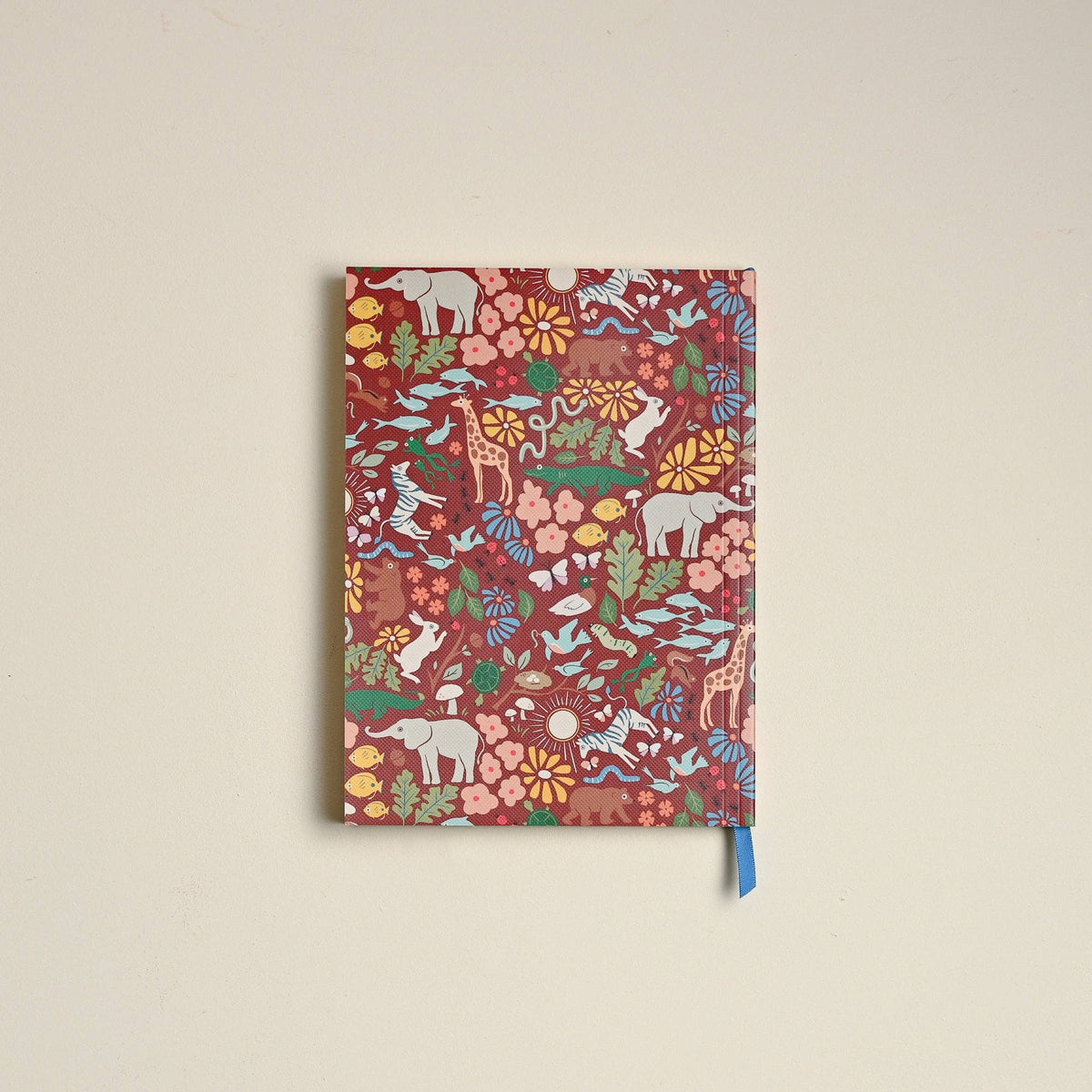 A hardcover notebook with a detailed safari-themed pattern featuring animals like elephants, giraffes, and lions, set against a warm red background with a blue ribbon bookmark, displayed on a light beige wall.