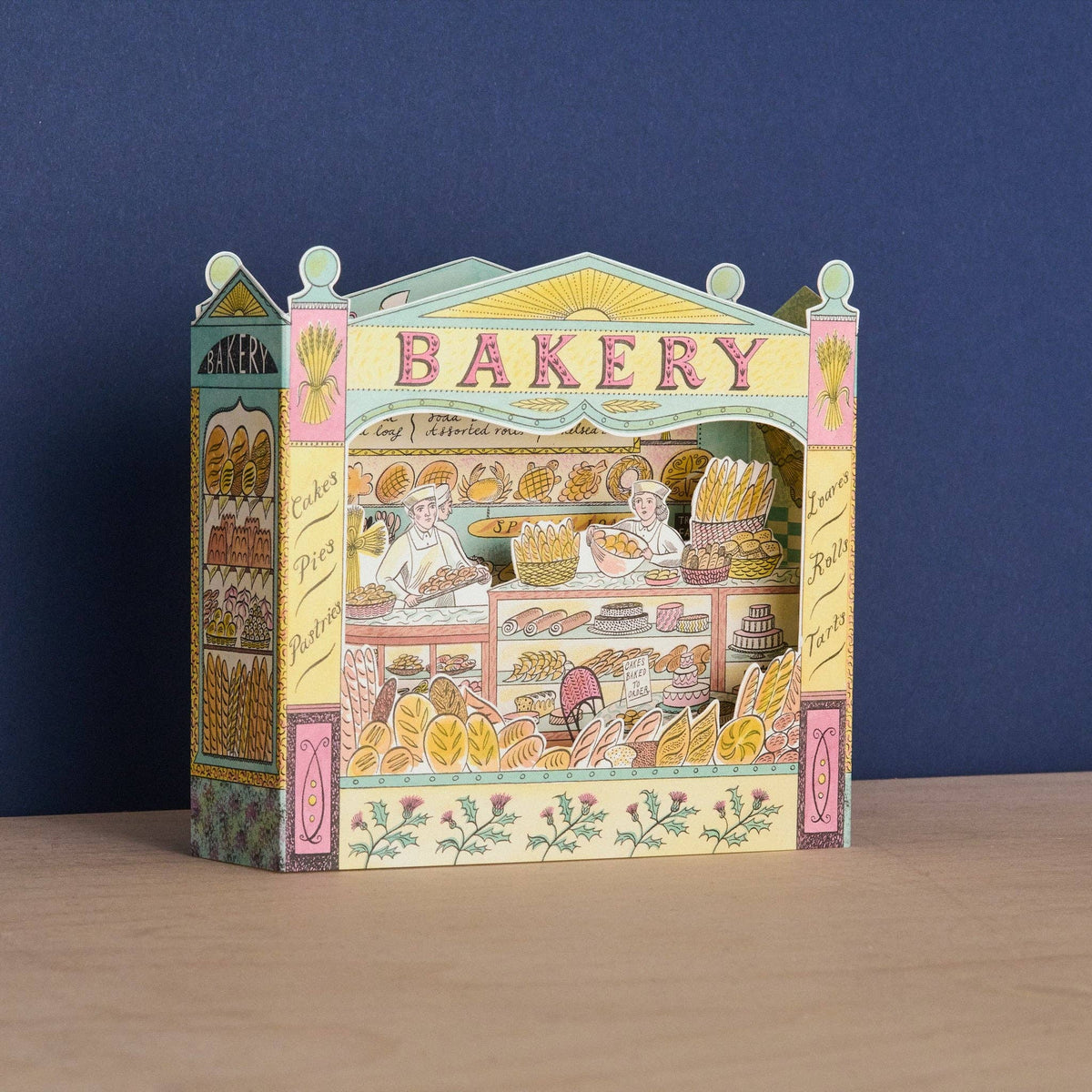 A colorful 3D papercraft model of a vintage bakery storefront, displayed on a light wooden surface against a dark blue background.