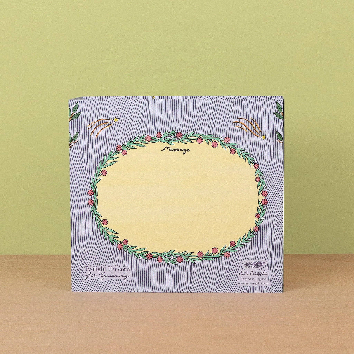 A square art print or card with a pale yellow center surrounded by a green wreath of red berries, set against a blue-and-white striped border. It is displayed on a light wooden surface in front of a pale yellow-green wall.