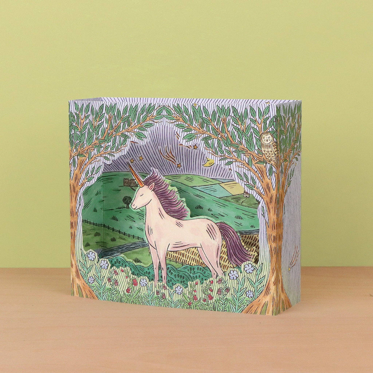A square decorative box with an illustrated fantasy scene of a unicorn in a grassy field framed by trees, set against a light olive green background and a pale wood surface.