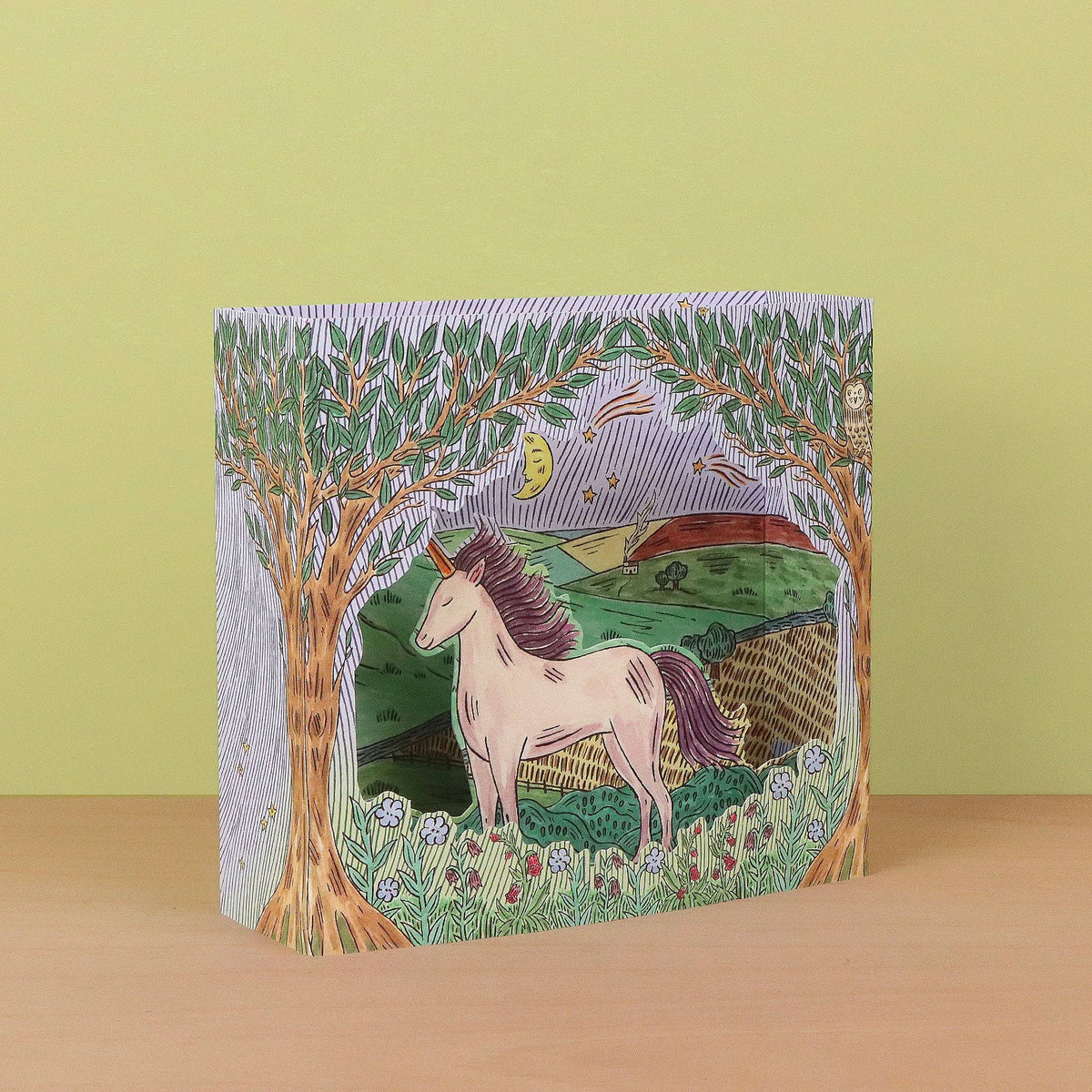 A rectangular decorative box standing upright on a wooden surface against a pale yellow-green background. The box is illustrated with a whimsical woodland scene featuring a white unicorn with a purple mane, trees, flowers, hills, and a crescent moon.