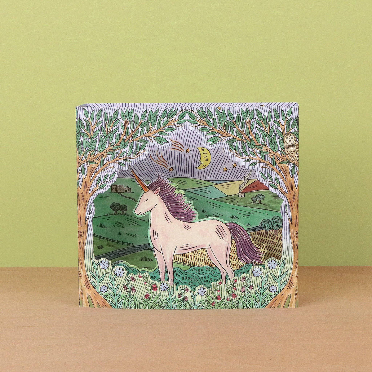 A square decorative card or art print featuring a whimsical illustrated unicorn in a pastoral landscape, standing upright on a light wood surface against a pale yellow-green background.