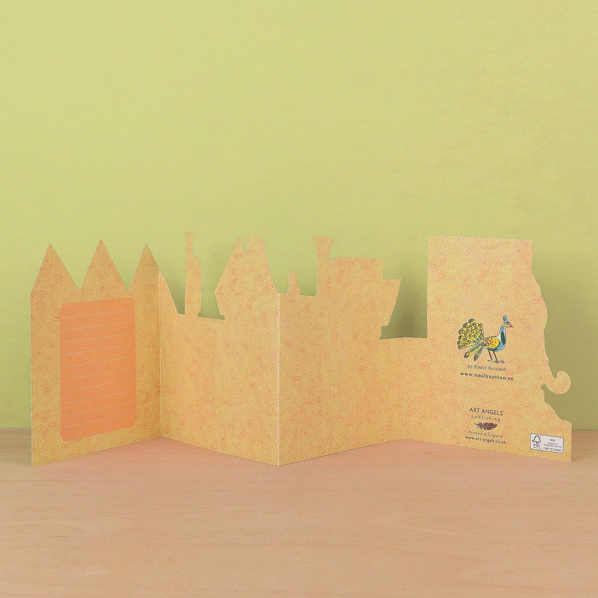 A handmade origami card or book made from textured light brown paper, standing open on a wooden surface against a pale yellow-green background. The left panel has a rectangular orange insert, and the right panel features a colorful peacock illustration with text.