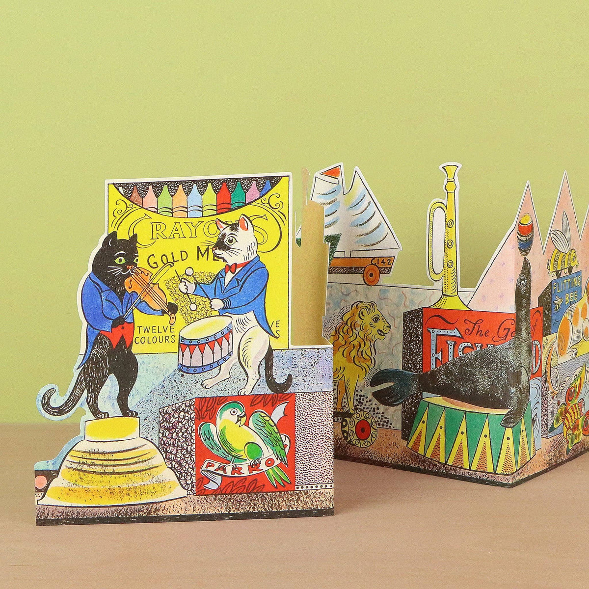 A colorful vintage-style pop-up cardboard display featuring circus and theatrical scenes, including a black cat playing an instrument, a lion, a parrot, and stage elements, set against a pale green background.
