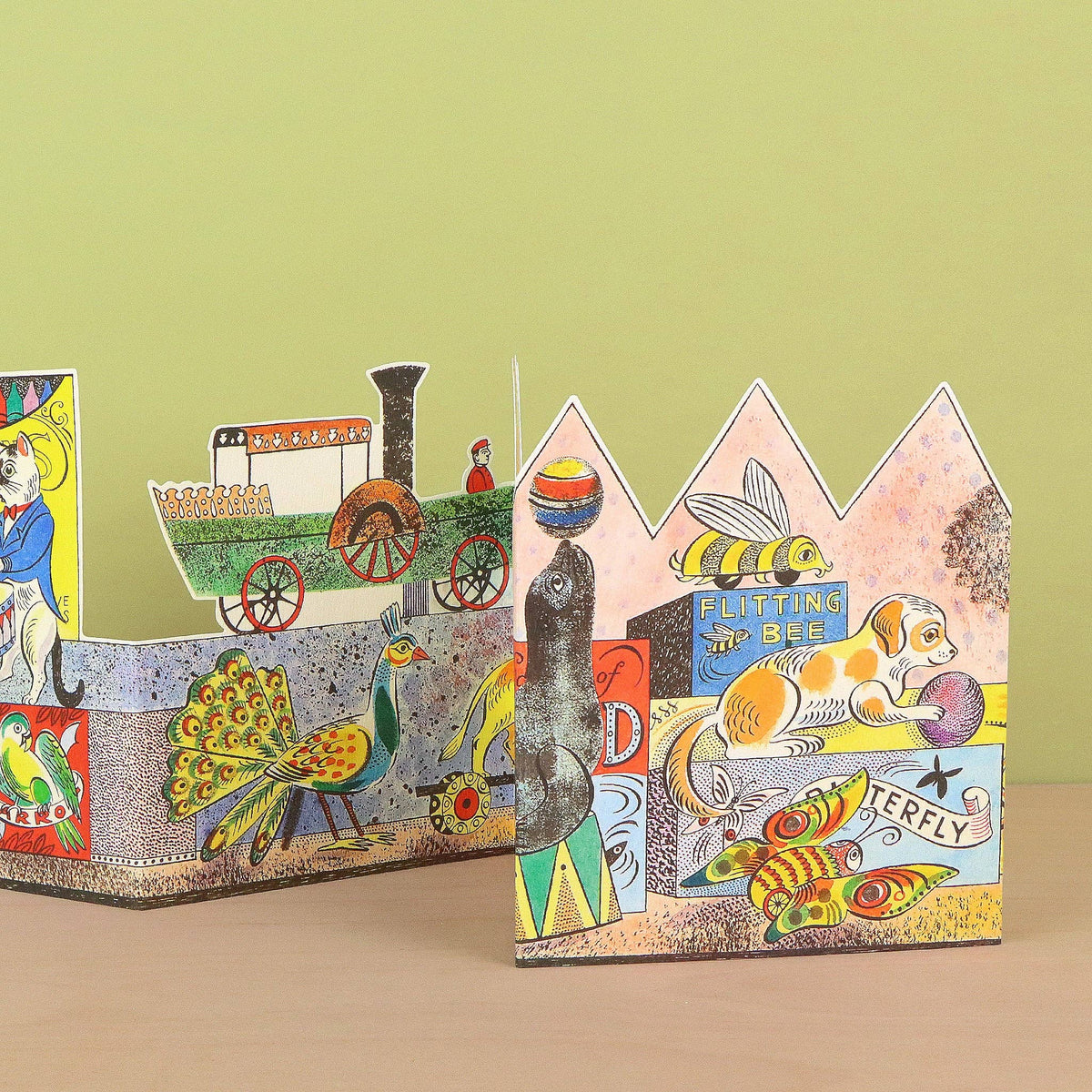 A set of colorful, handcrafted cardboard pop-up toys featuring whimsical illustrations of a steam train, a peacock, a bee, a dog, and a fish, displayed on a light wood surface against a solid light green background.