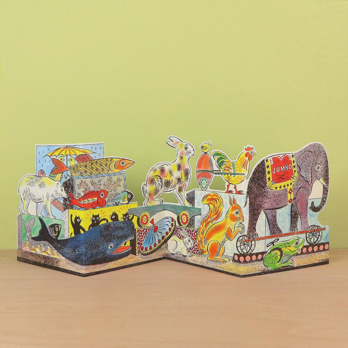 A colorful 3D pop-up cardboard animal storybook displayed on a light wooden surface against a pale yellow-green background. The pop-up features various animals including an elephant, rabbit, rooster, squirrel, whale, lion, and fish, each with vibrant painted details and traditional patterns.