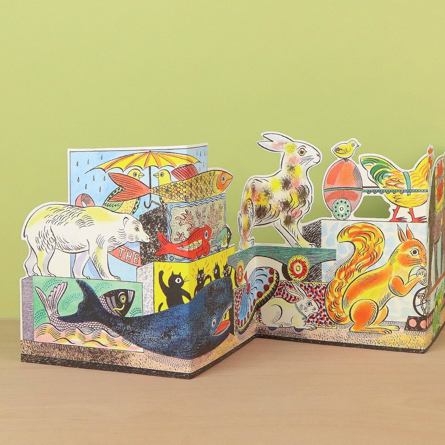 A colorful 3D pop-up cardboard animal storybook displayed on a light wooden surface against a pale green background. The book features various illustrated animals including a white polar bear, a blue whale, a red fox, a yellow squirrel, a rabbit, a fish, a chicken, and a rooster.