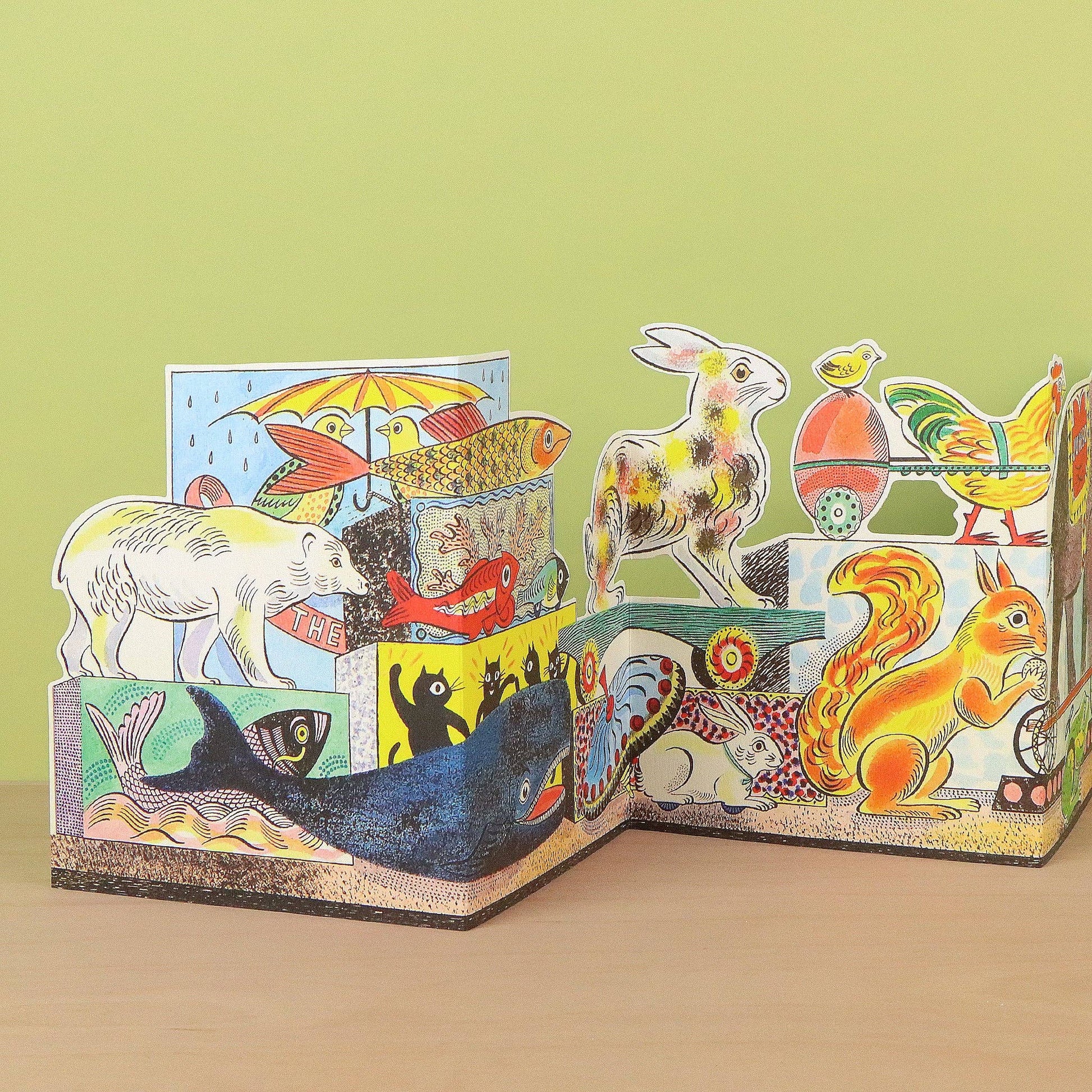 A colorful 3D pop-up cardboard animal storybook displayed on a light wooden surface against a pale green background. The book features various illustrated animals including a white polar bear, a blue whale, a red fox, a yellow squirrel, a rabbit, a fish, a chicken, and a rooster.