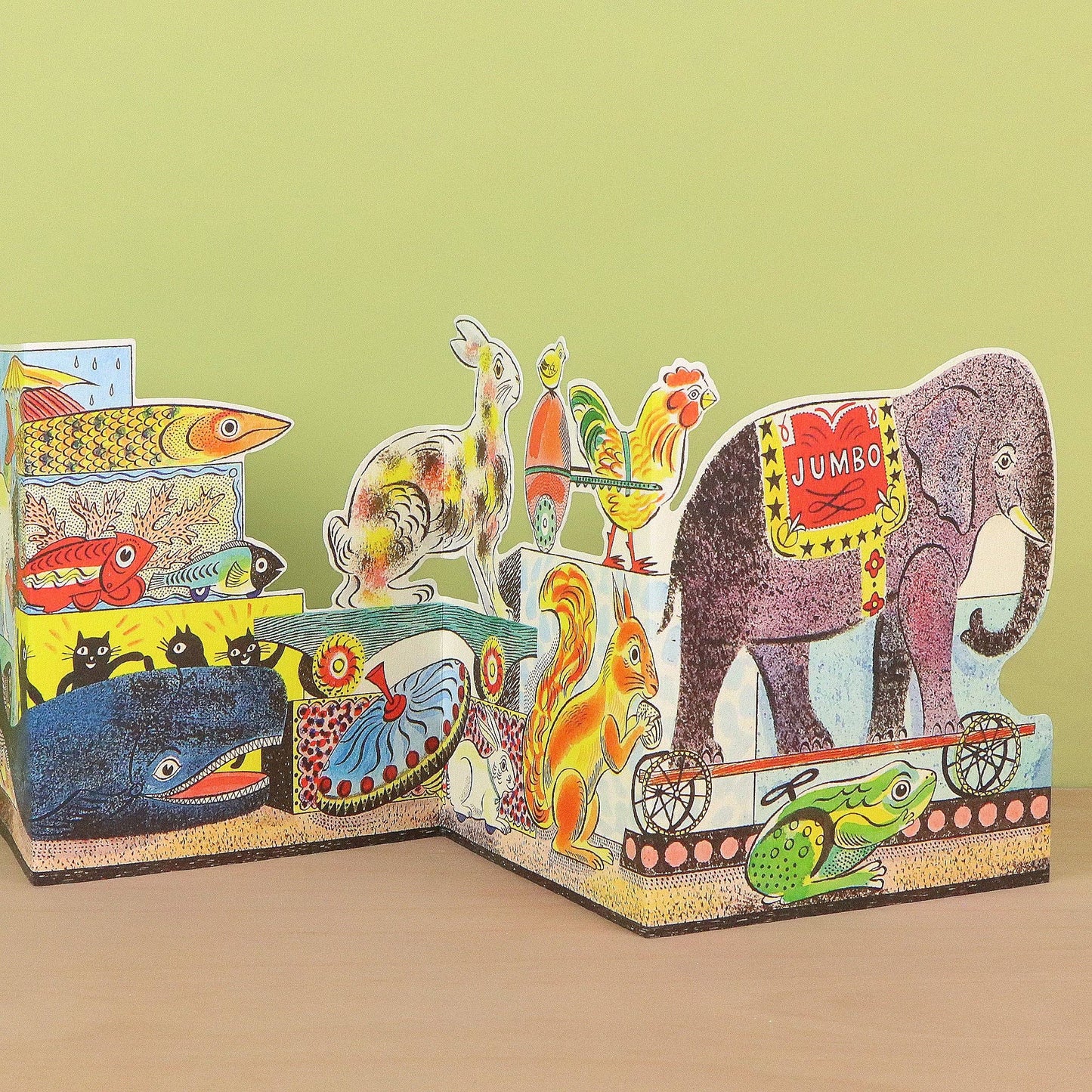 A colorful children's wooden animal storybook with tiered pages showing illustrated animals including an elephant, fish, whale, rabbit, rooster, squirrel, frog, and a horse, displayed on a tabletop against a pale green background.