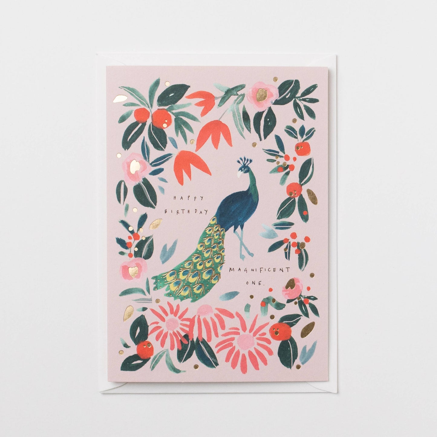 A square greeting card with a light pink background featuring an illustrated peacock surrounded by flowers and leaves, shown with a white envelope on a white background.