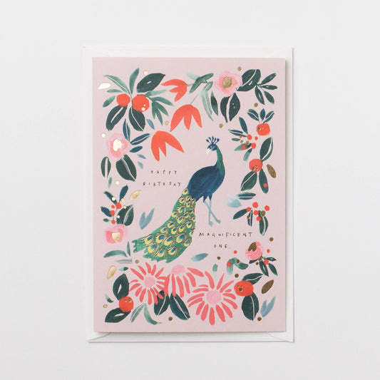 A square greeting card with a light pink background featuring an illustrated peacock surrounded by flowers and leaves, shown with a white envelope on a white background.