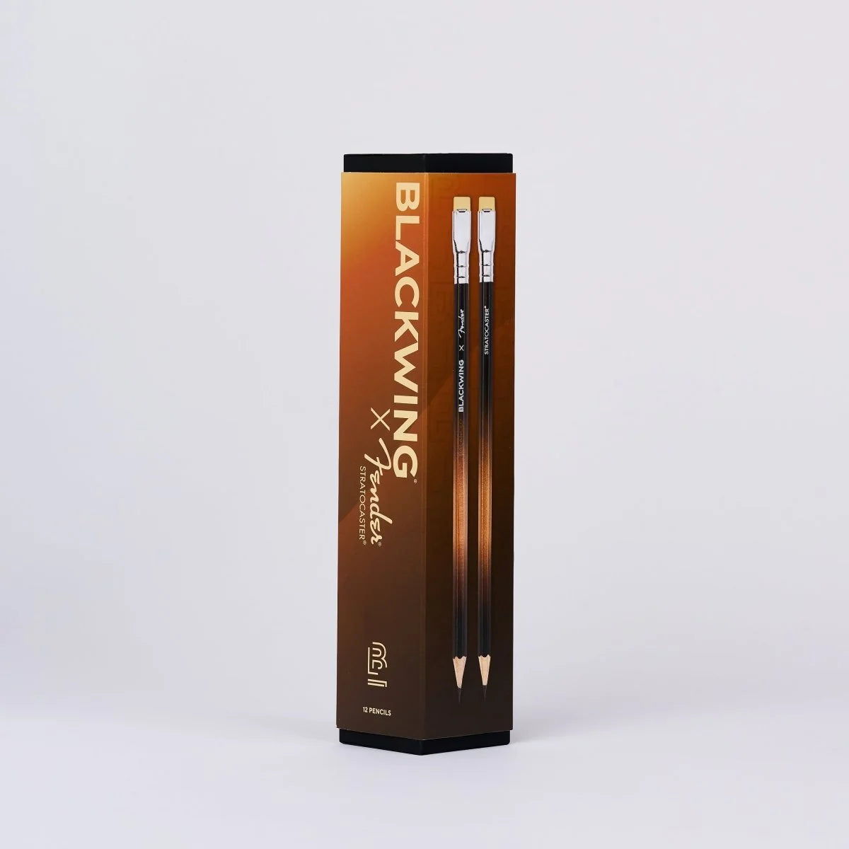 A tall rectangular product box for Kuretake Blackwing x Graphite pencils, standing upright on a light gray studio background. The box is dark brown and black with gold gradient text and shows two pencils inside.