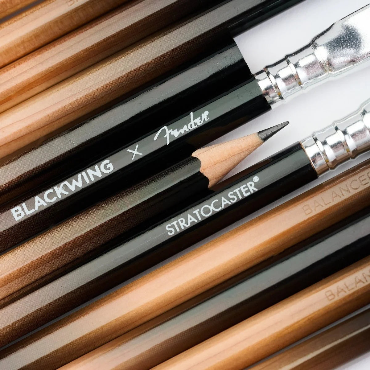 Close-up of several Fender Blackwing Stratocaster pencils arranged diagonally, showing black ferrules, silver metal tips, and natural wood barrels with white printed branding.
